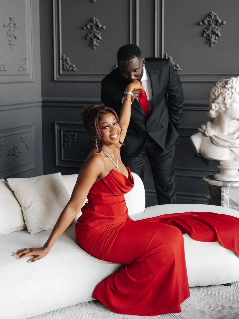 Harrison kissing Otobong’s hand as her red dress falls across the sofa